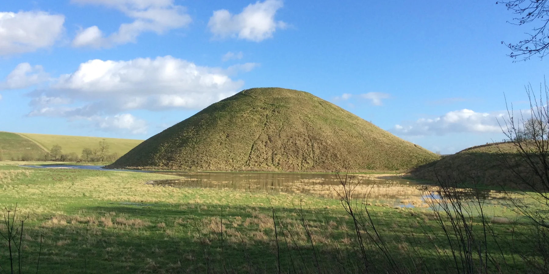 Silbury Hill, a conical grass-covered prehistoric mound, reflected in a pond with chalk downland behind